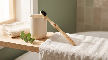 A bamboo toothbrush with a black bristle head leaning against a cream ceramic cup on a light wood shelf beside a folded washcloth, a linen towel and a sprig of green leaves in soft window light