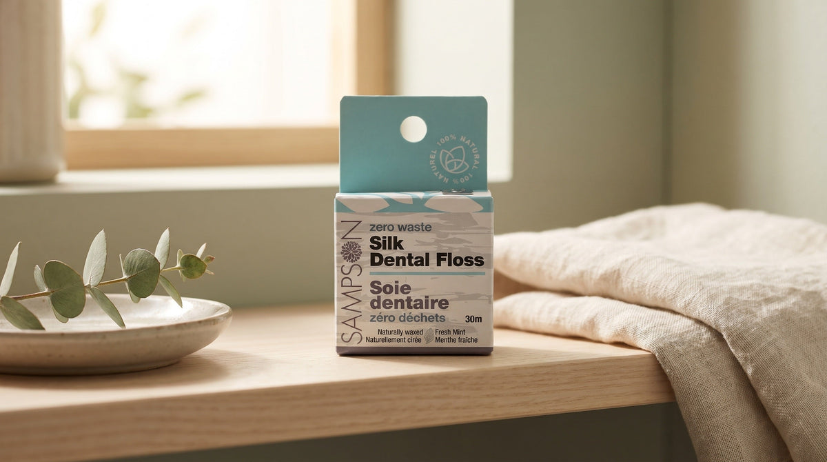 Sampson Silk Dental Floss box with its teal hanging card on a pale wood bathroom shelf beside a folded linen towel and a eucalyptus sprig on a ceramic dish in soft window light