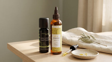 A Sampson jojoba oil bottle beside a Sampson essential oil bottle on a pale wood table, with a glass dropper resting on a speckled ceramic dish and dried sage on folded cream linen, in soft window light