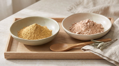Two ceramic bowls of clay powder, golden multani mitti and pale rhassoul, on a wooden tray