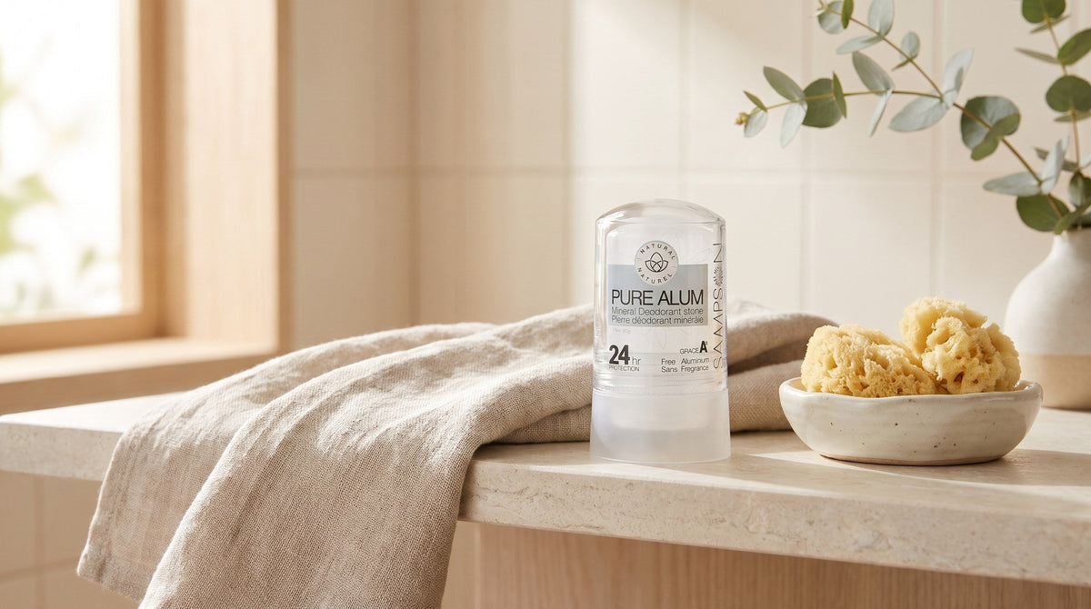 Clear Sampson Pure Alum mineral deodorant stone on a pale stone bathroom shelf beside a folded linen towel, a natural sea sponge in a ceramic dish and eucalyptus in soft window light