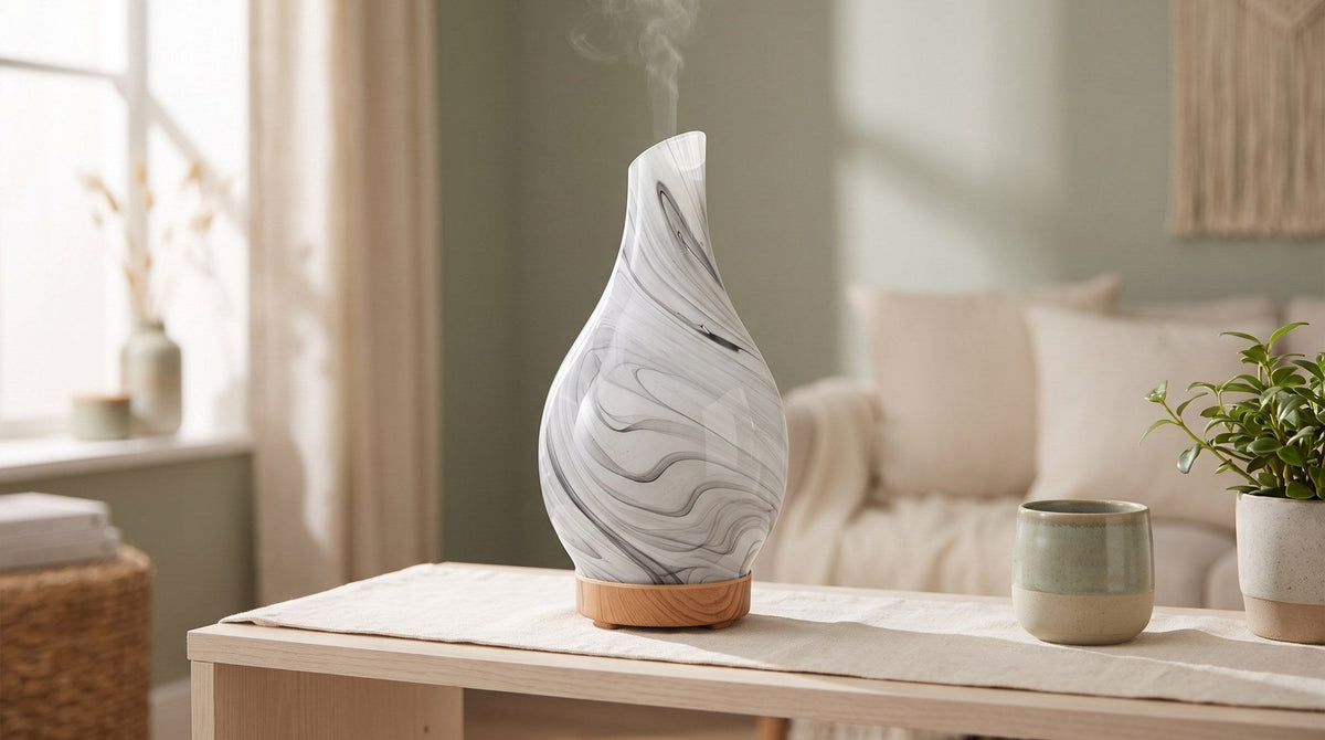 A marbled grey and white glass aroma diffuser with a wooden base releasing a faint mist on a pale wood table beside a ceramic cup and a small green plant in a sunlit living room
