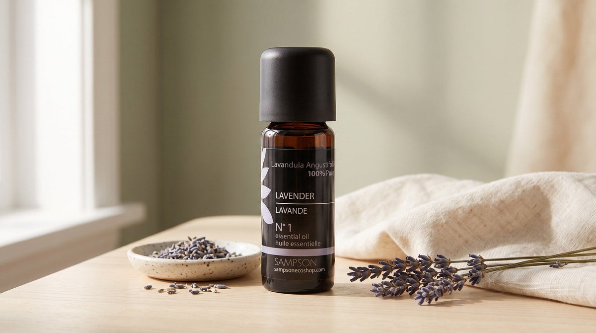 A Sampson lavender essential oil bottle on a pale wood table beside a speckled ceramic dish with dried lavender sprigs and folded cream linen, in soft window light