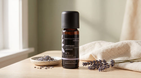 A Sampson lavender essential oil bottle on a pale wood table beside a speckled ceramic dish with dried lavender sprigs and folded cream linen, in soft window light