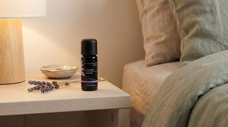 A Sampson lavender essential oil bottle on a pale wood bedside table beside dried lavender stems, a small ceramic dish and a glowing lamp, with linen bedding at the edge of the frame