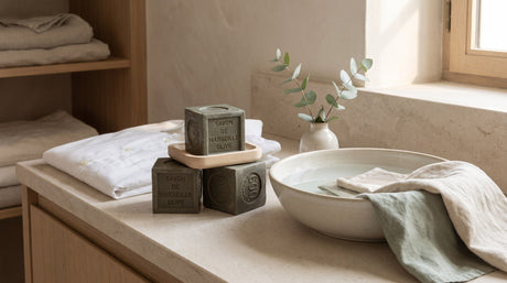 Traditional olive oil Marseille soap cubes on a wooden dish beside a folded white shirt, a ceramic basin of water and a linen cloth on a stone counter