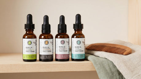 Four amber dropper bottles of Sampson beard oil on a pale wooden shelf beside a wooden beard comb and a folded linen towel in soft natural light