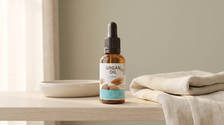Amber glass dropper bottle of Sampson argan oil on a pale wooden bathroom shelf beside a folded linen towel and a ceramic dish in soft window light