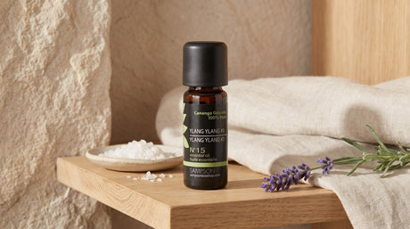 Sampson Ylang Ylang essential oil bottle with its SAMPSON label on a pale wood shelf beside a ceramic dish of salt, a folded linen towel and a sprig of lavender in soft window light