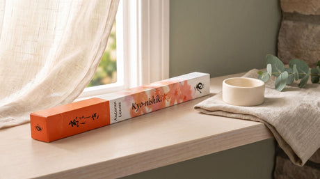 A slim orange and white Japanese incense box lying on a pale wood windowsill beside a small cream ceramic holder and a folded linen cloth, with a sheer linen curtain and an open window in soft daylight