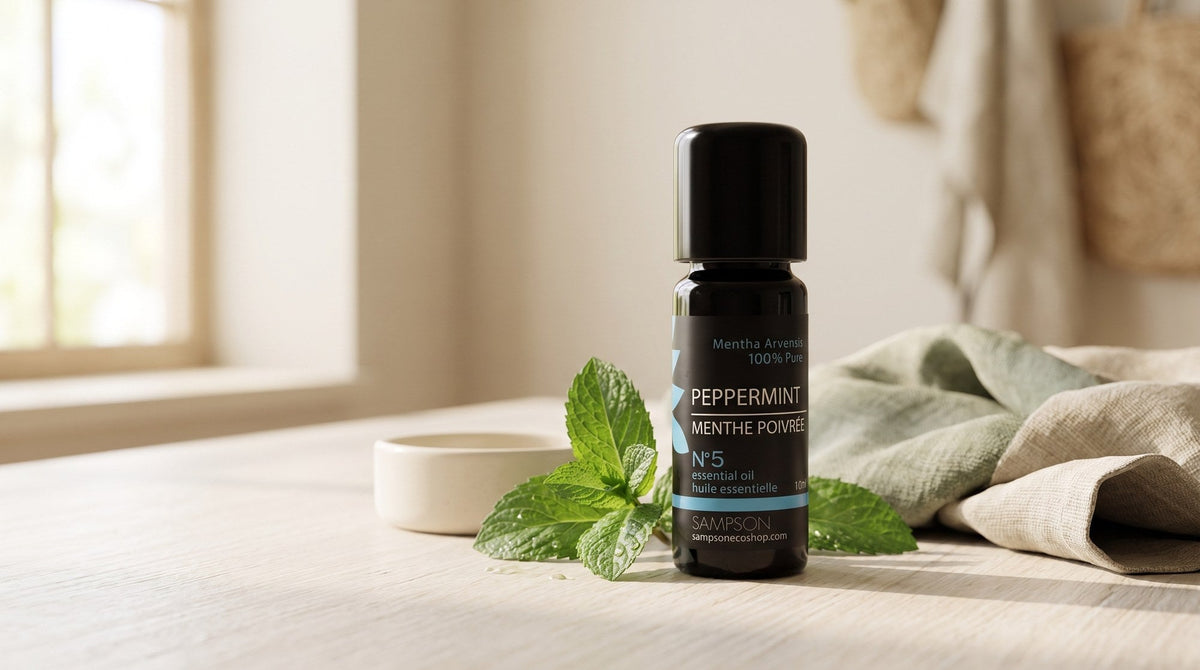 A Sampson peppermint essential oil bottle on a pale wood table beside fresh peppermint leaves, a small ceramic dish and folded linen, in soft window light