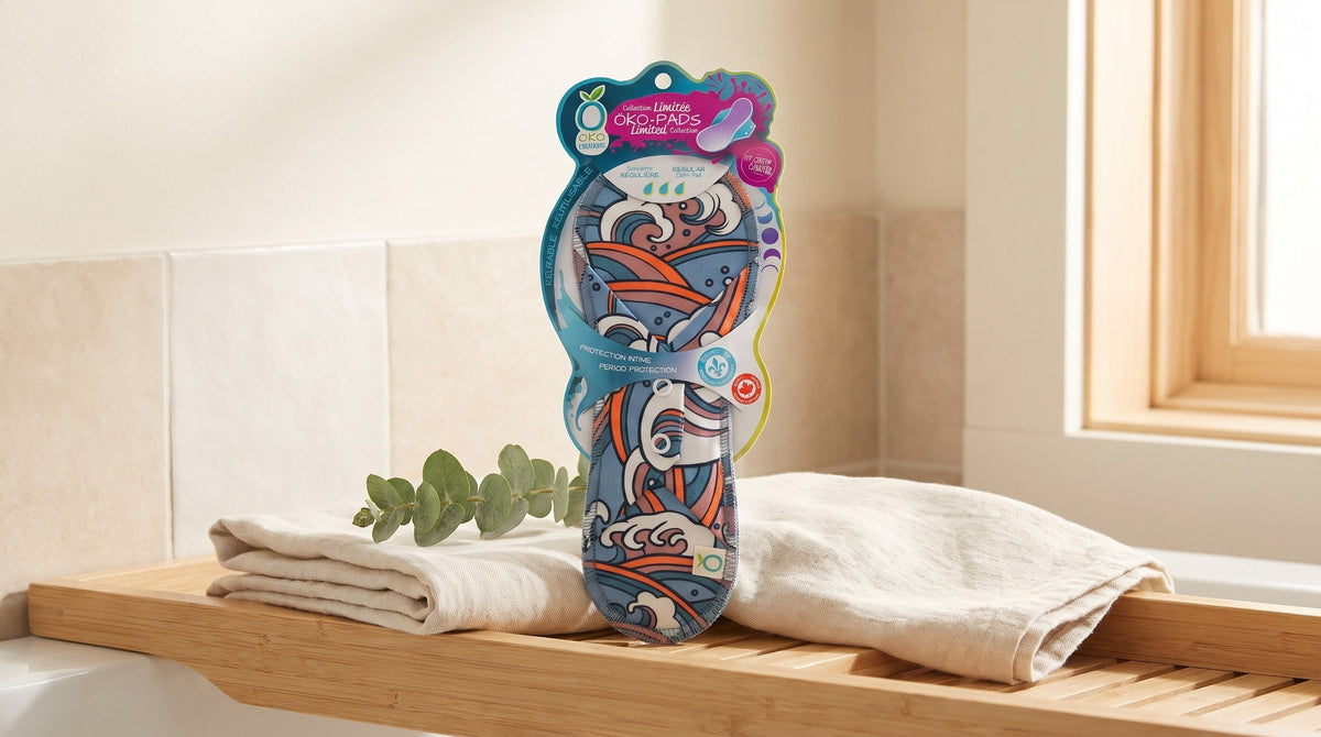 OKO reusable menstrual pad in its retail card packaging standing on a pale wood bath shelf beside a folded linen towel and a eucalyptus sprig in soft window light