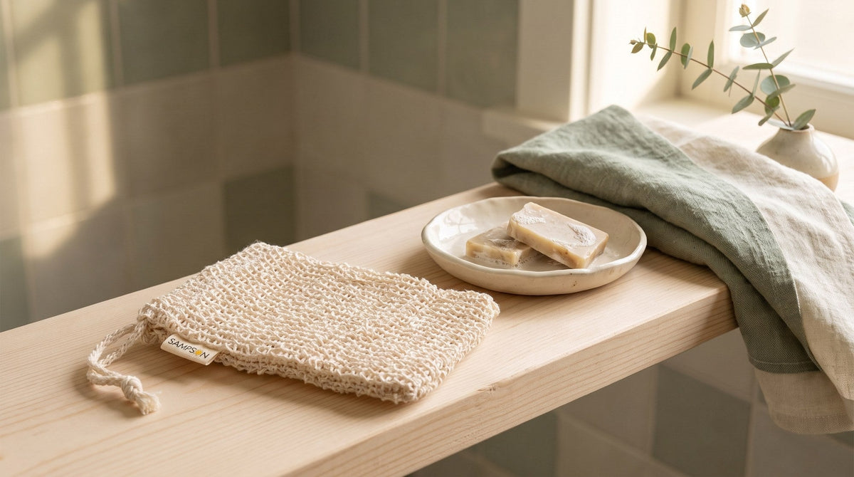 A natural sisal soap saver bag with a Sampson label on a pale wood bathroom shelf beside a small dish of soap slivers and a folded linen towel